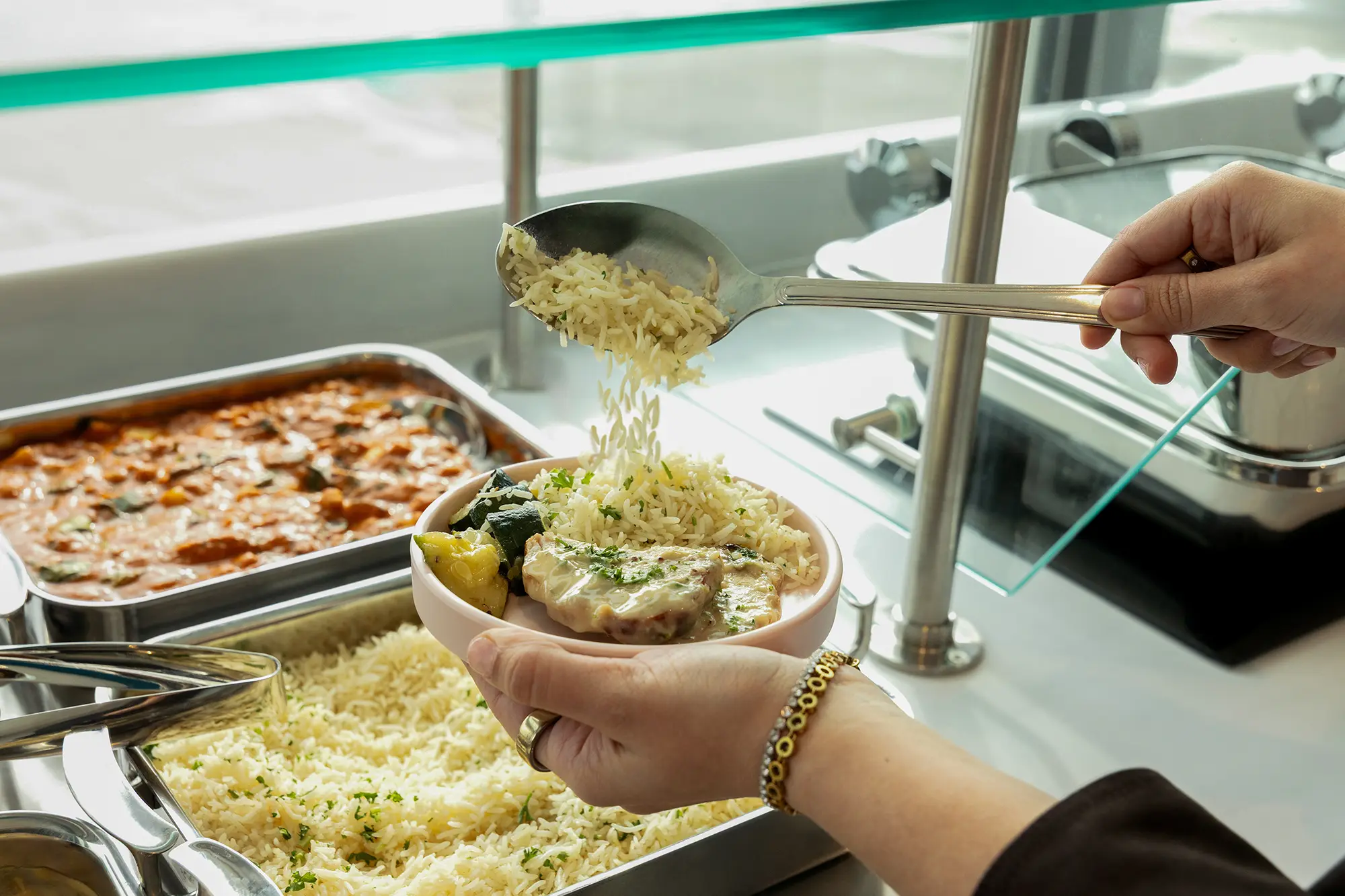 A hand spoons herbed basmati rice onto a bowl of chicken and vegetables at a hotel buffet, with trays of tomato-based stew and rice visible in the background.