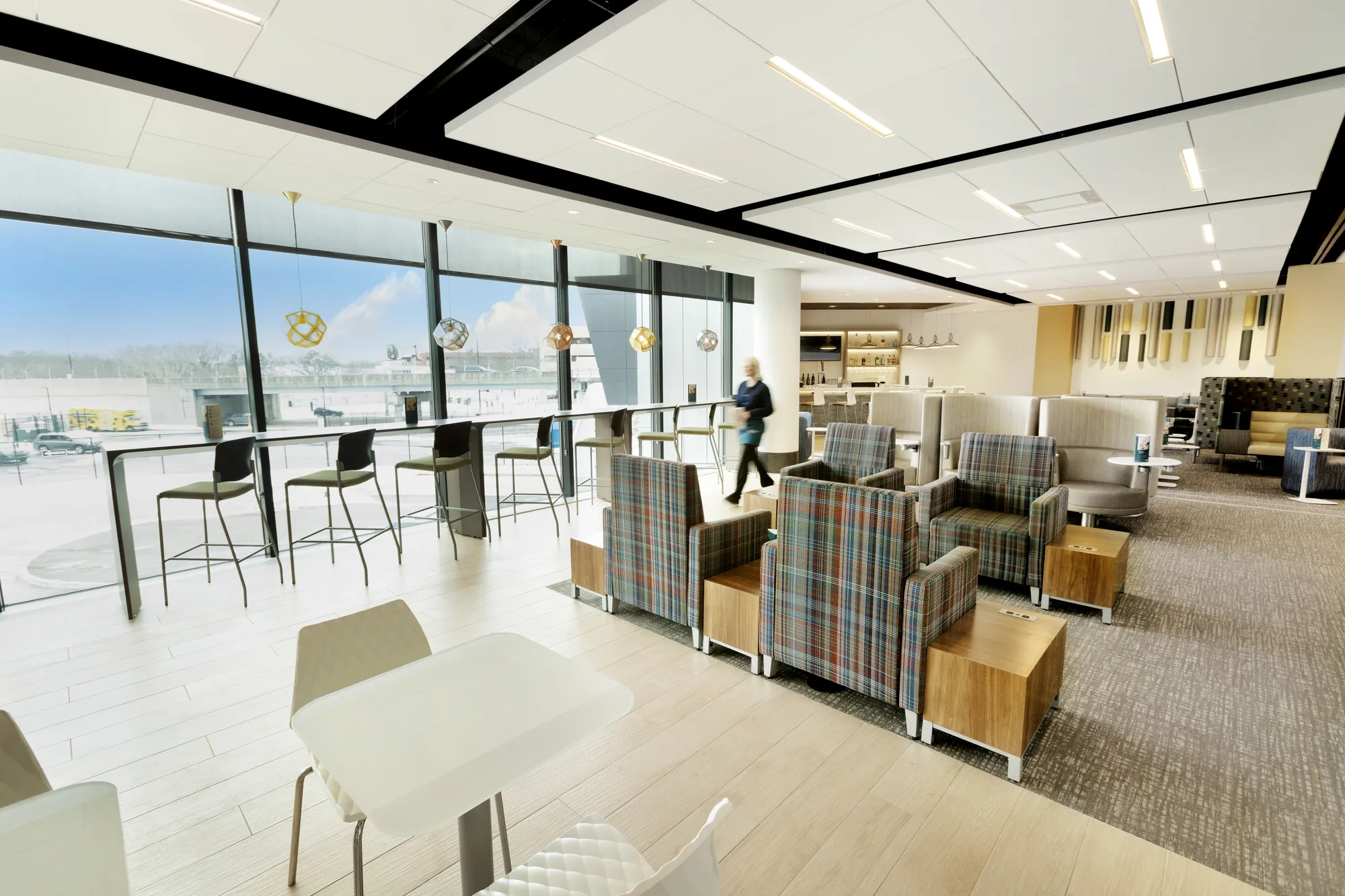 Interior of Escape Lounge at Bradley International Airport, featuring plaid armchairs, high-top seating along floor-to-ceiling windows overlooking the airfield, and a stocked bar in the background.