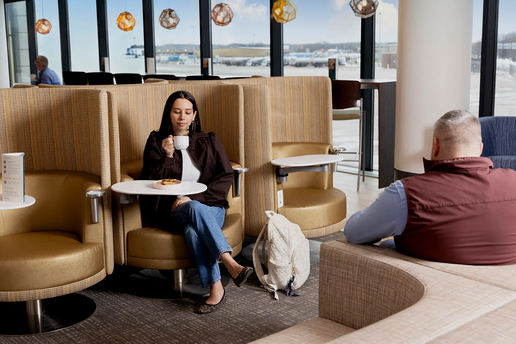 A woman sips coffee while relaxing in a high-backed swivel chair with a personal side table in an airport lounge, with hanging geometric lights and tarmac views through floor-to-ceiling windows behind her.